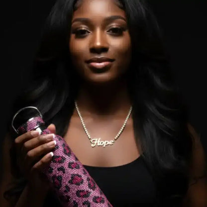 A dark-skinned woman with long, dark hair, wearing a black top and a ’Hope’ necklace, holding a pink leopard print water bottle.