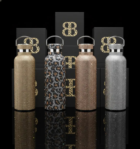 Four sparkling, rhinestone-encrusted water bottles in gold, leopard print, rose gold, and silver, each with a sleek metallic handle.