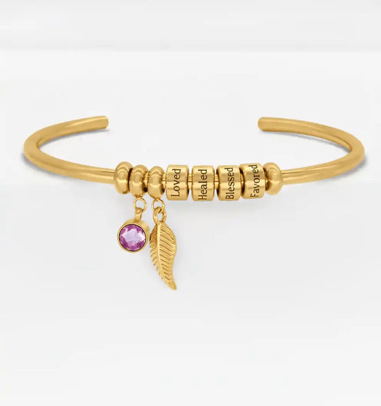 Gold cuff bracelet with engraved beads, a purple gemstone, and a feather charm.