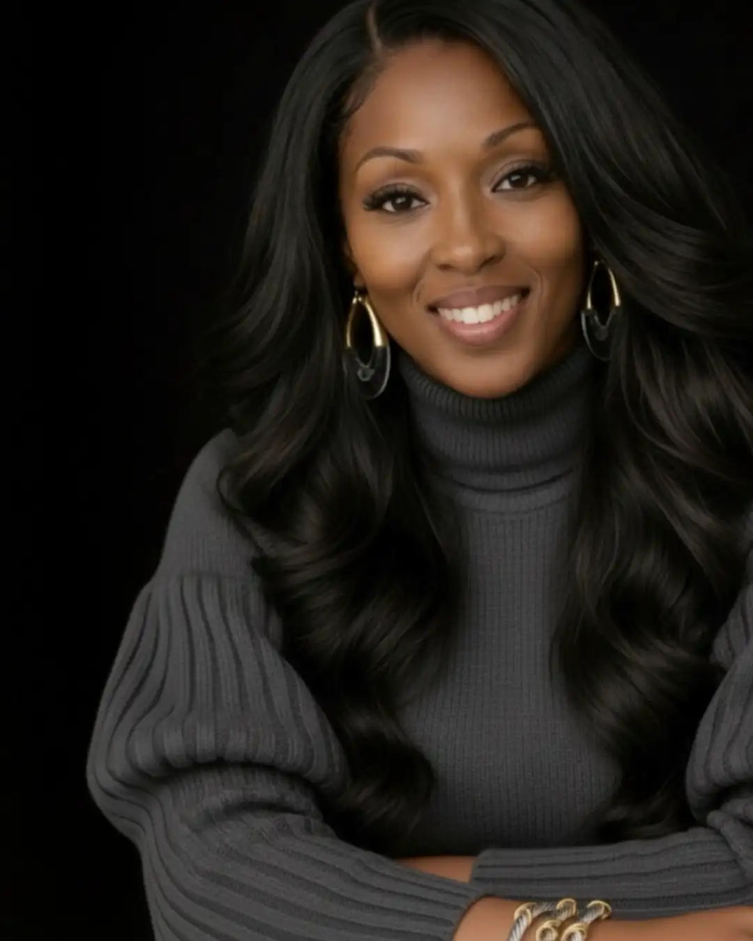 A woman wearing a dark gray ribbed turtleneck sweater with long, flowing black hair and gold hoop earrings.