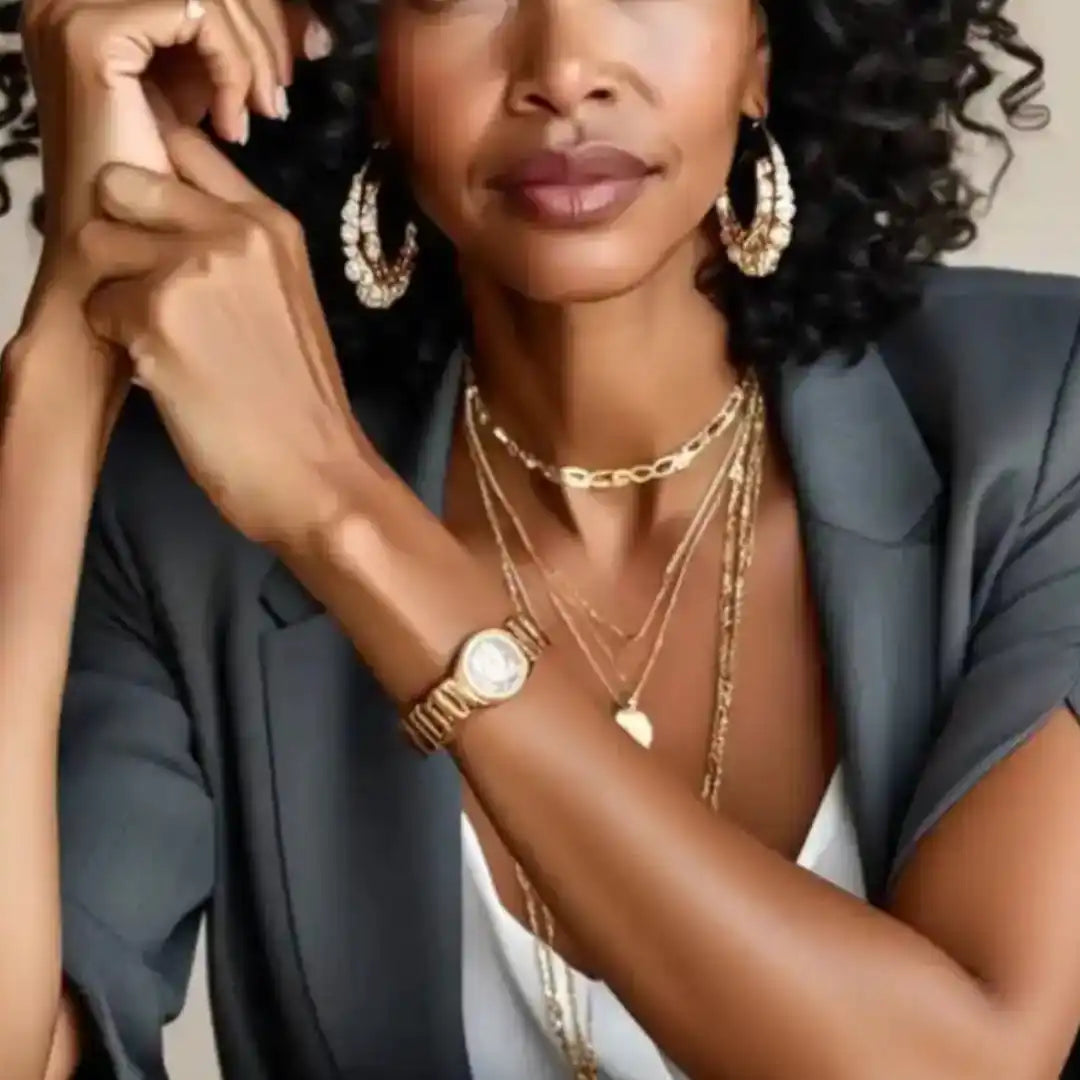 A woman wearing a gray blazer, a white top, and layered gold necklaces, with a gold watch on her wrist and large gold hoop earrings.